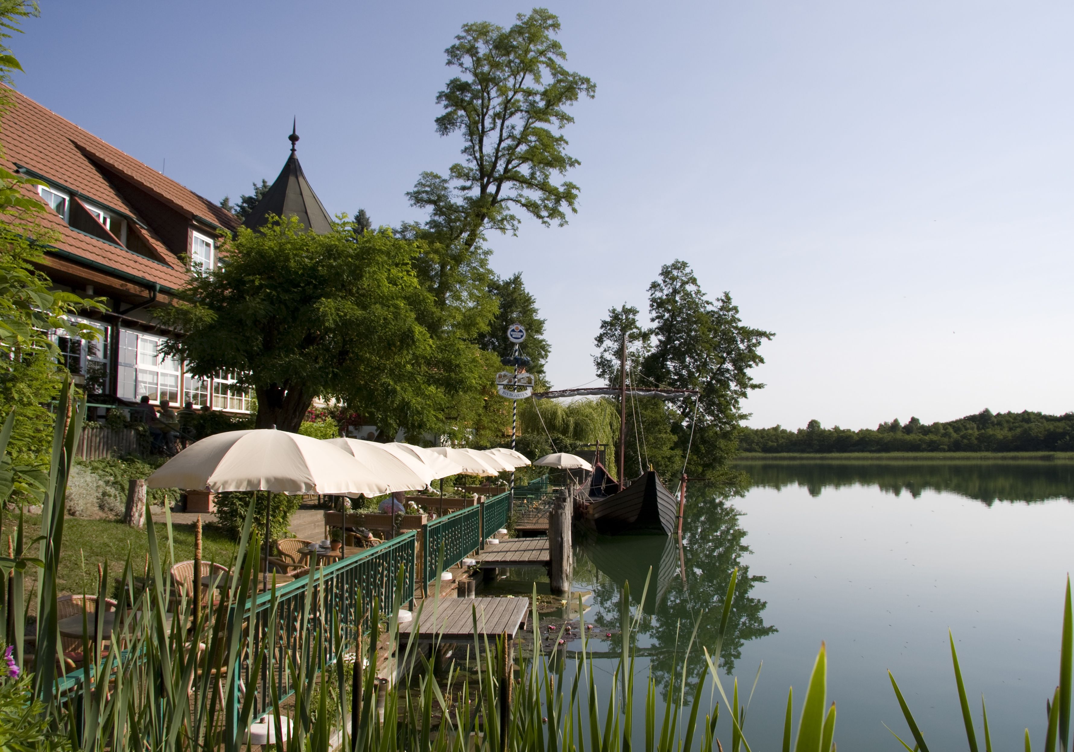 A riverside restaurant with outdoor seating, a wooden dock, and a bridge in the background.