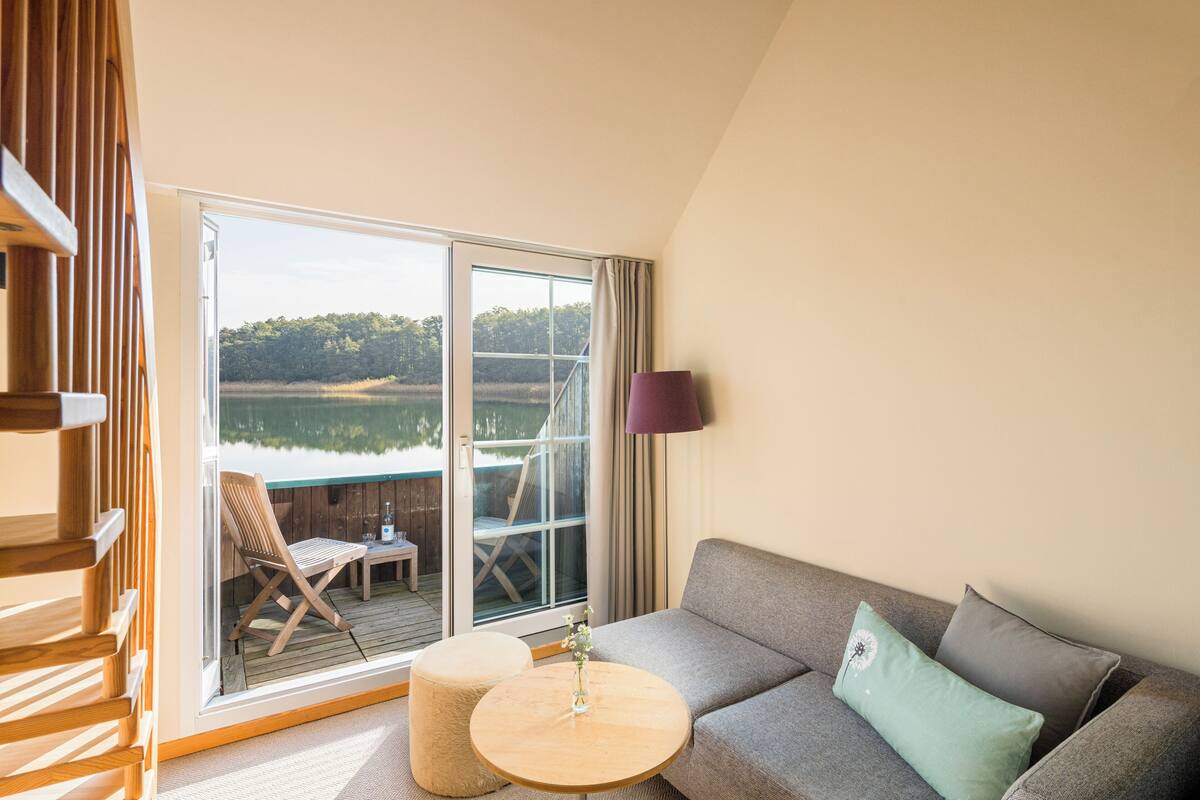Une pièce comprenant un canapé, un pouf et une petite table sur laquelle est posé un vase. Une fenêtre offre une vue sur un lac et des arbres.