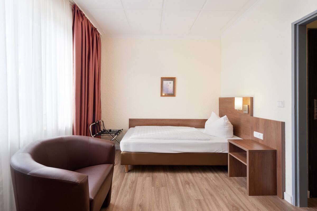 A hotel room with a bed, a chair, a bedside table, and a wall-mounted lamp.
