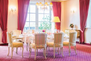 A dining room with a round table set for four, surrounded by striped chairs, with a chandelier above and a window with curtains. - Hotel Landhaus Höpen (Schneverdingen)