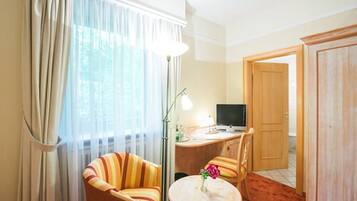 A hotel room with a bed, a desk with a computer, a chair, a small table with a vase, and a window with curtains.
