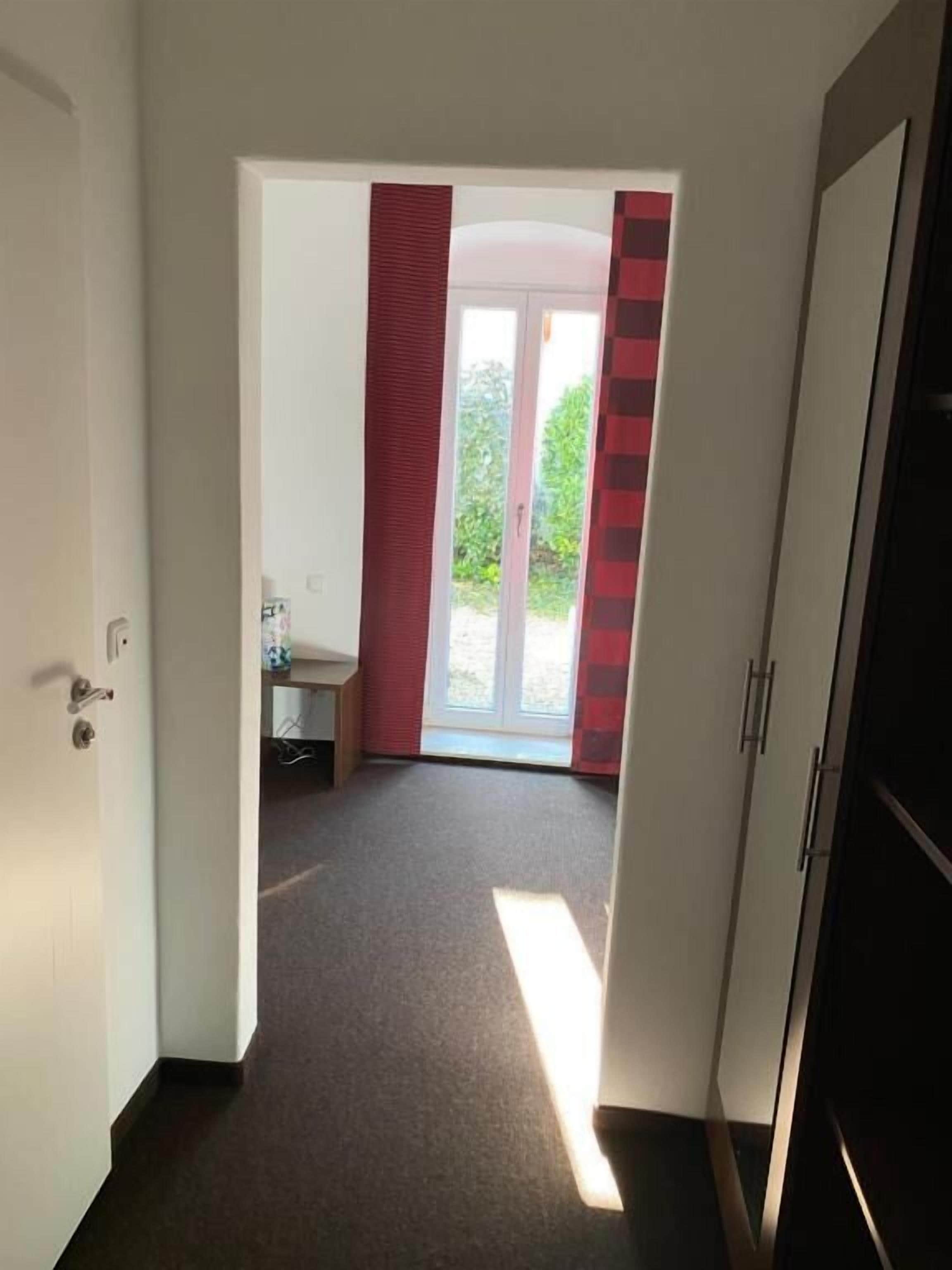 A hallway leading to a room with a door, a window, and a view of greenery outside.