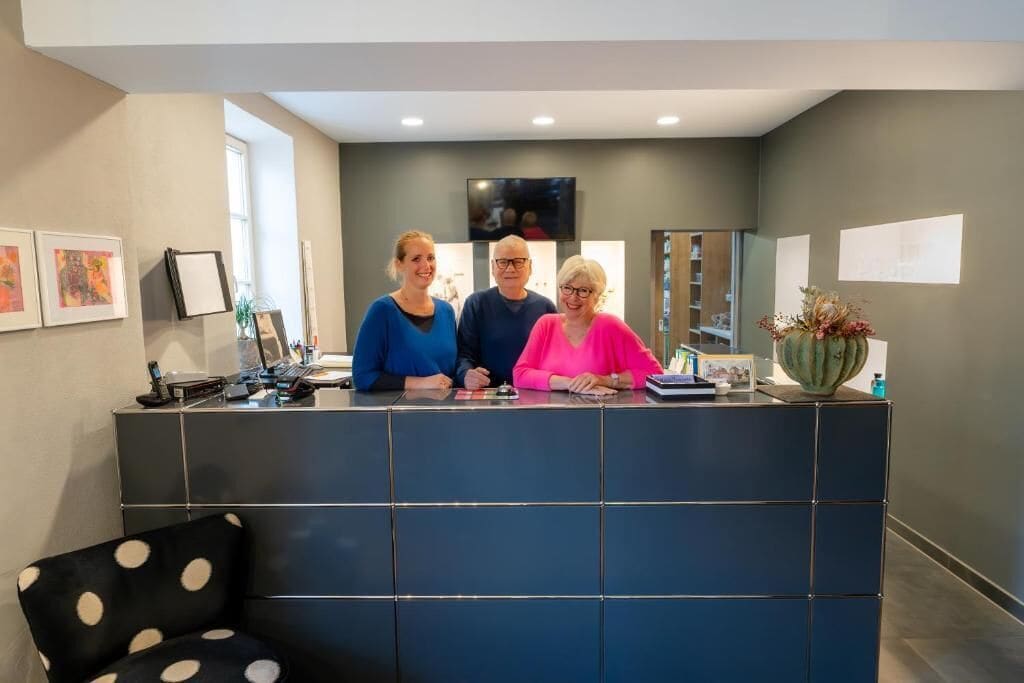 A reception desk with three individuals standing behind it, a television mounted on the wall, and a vase with flowers on the counter.