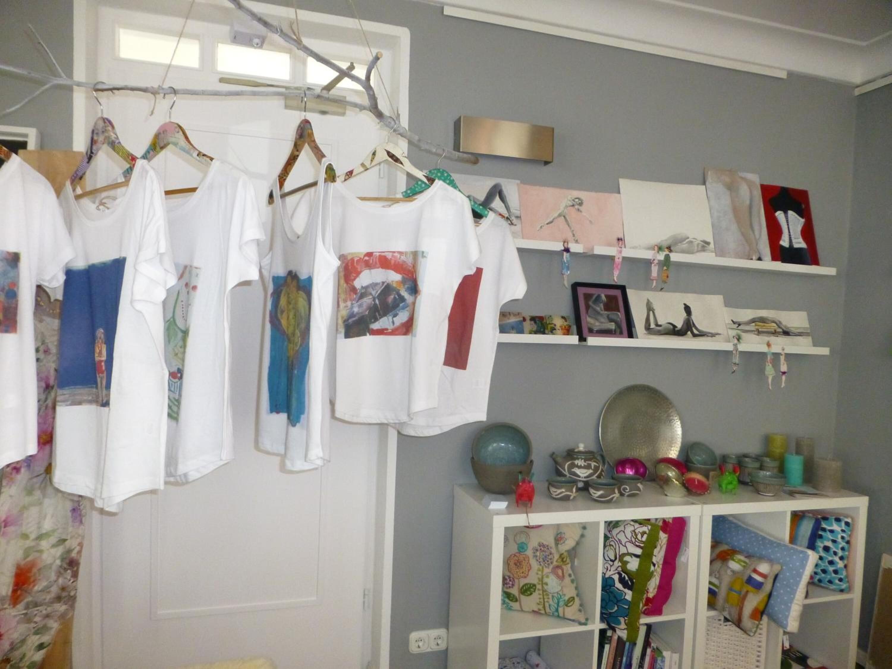 A room with a white wardrobe, a clothing rack with various t-shirts, and shelves with decorative items and artwork.