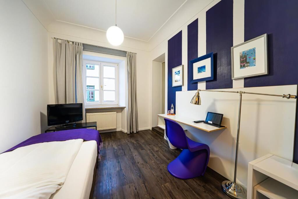 Una habitación de hotel con una cama, un escritorio con una computadora portátil, una silla, un televisor y una ventana con cortinas.
