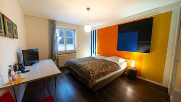 A hotel room with a bed, a desk with a computer, a window with curtains, and a wall with a geometric design.