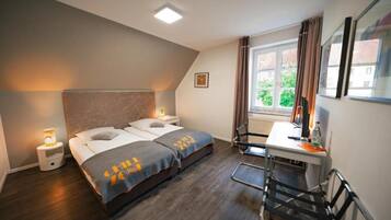 Una habitación de hotel con cama, mesitas de noche, escritorio, silla y ventana con cortinas.