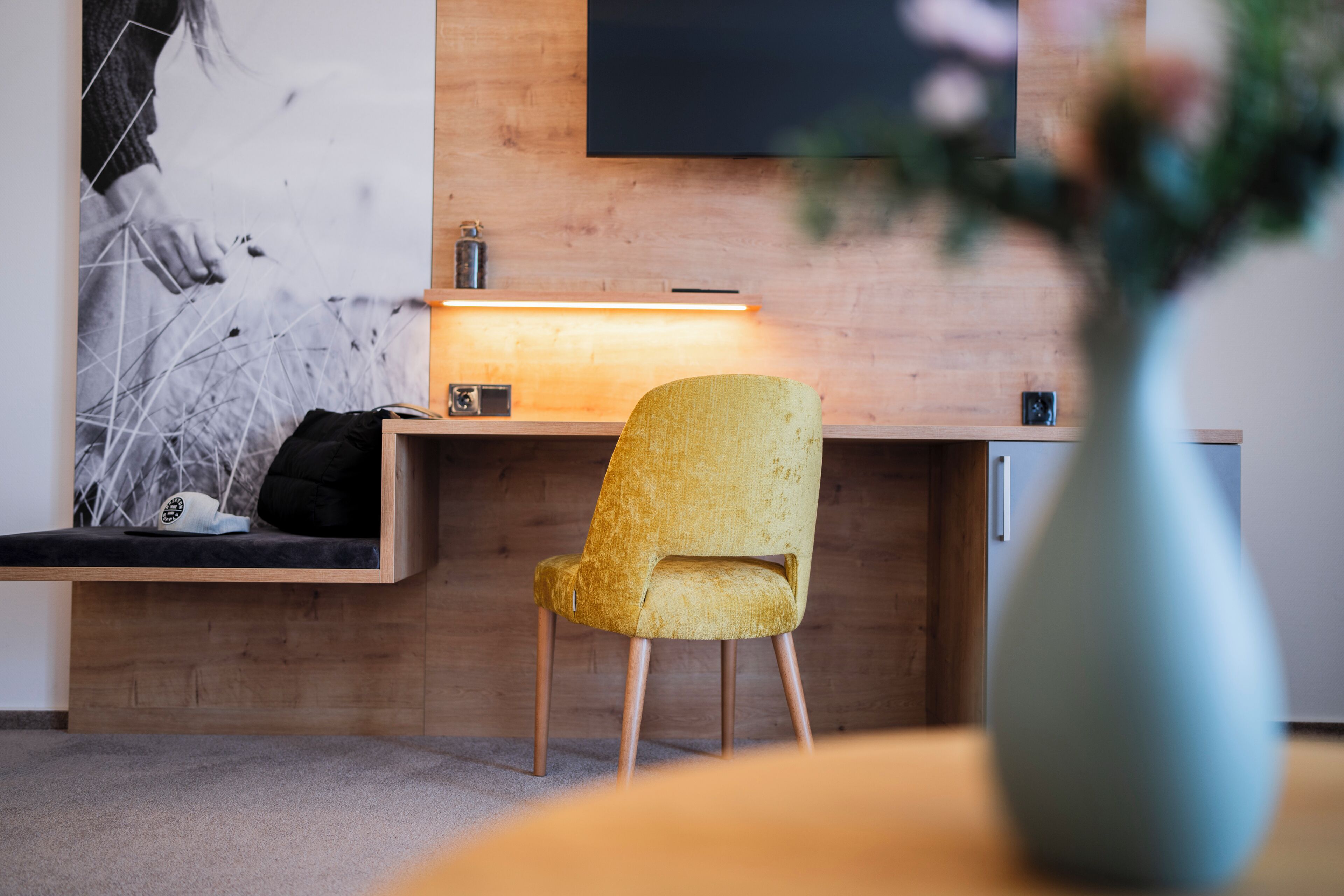 A modern room with a wooden desk, a yellow chair, a vase with flowers, and a wall-mounted TV.
