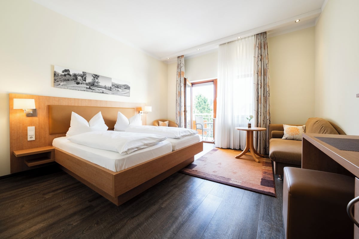 A modern hotel room with a large bed, a desk, a chair, and a balcony with curtains.