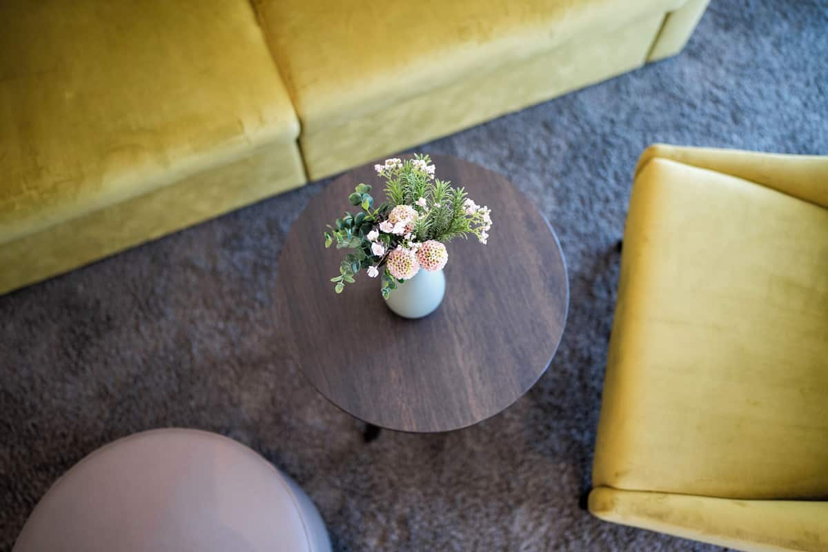 A round wooden table with a white vase containing pink flowers and green foliage.