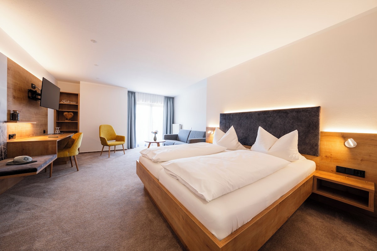 A modern hotel room with a large bed, a desk, a chair, a TV, and a bookshelf.