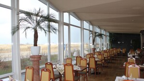 A dining area with tables and chairs arranged for service, large windows with a view of the outdoors, and a palm tree in the center. - Hotel Stolteraa (Rostock)