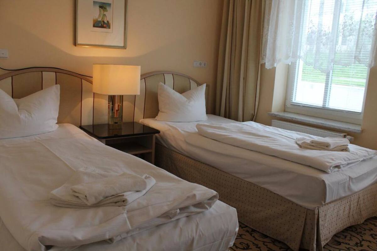 A hotel room with two single beds, a nightstand with a lamp, a window with curtains, and a framed picture on the wall.