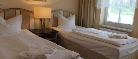 A hotel room with two single beds, a nightstand with a lamp, a window with curtains, and a framed picture on the wall.