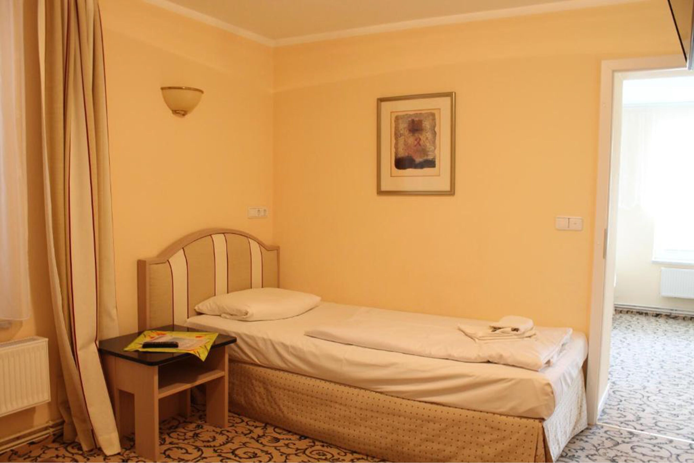 A small, single-bed room with a wooden headboard, a bedside table with a magazine, and a framed picture on the wall.