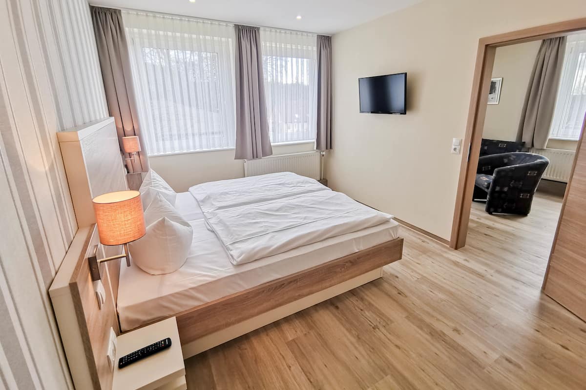 A modern hotel room with a wooden bed, a bedside table with a lamp, a TV mounted on the wall, and a view of the hallway.