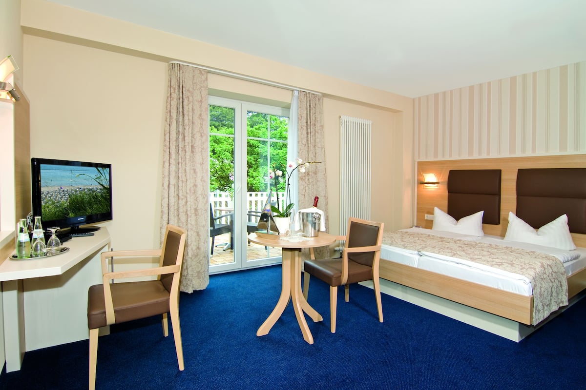 A hotel room with a bed, a desk with a TV, two chairs, a table with a lamp, and a view of greenery outside.