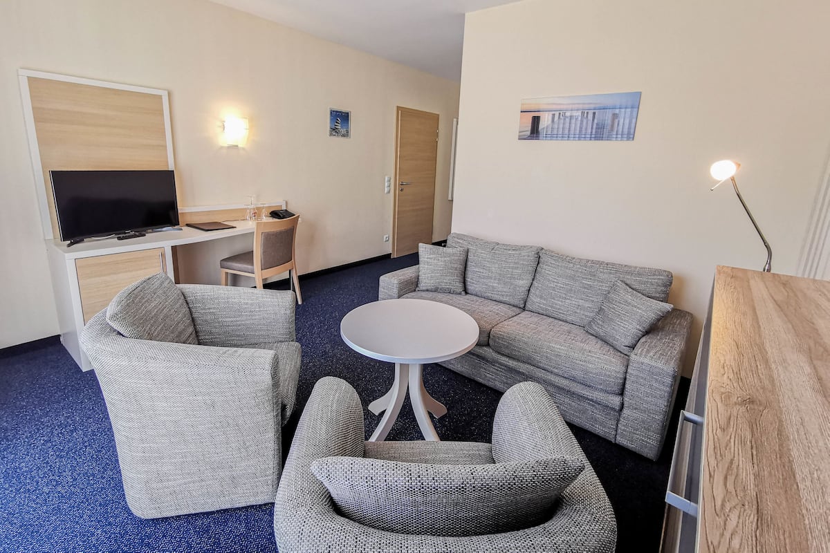 A hotel room with a sofa, two armchairs, a small round table, a TV, a desk, and a door.