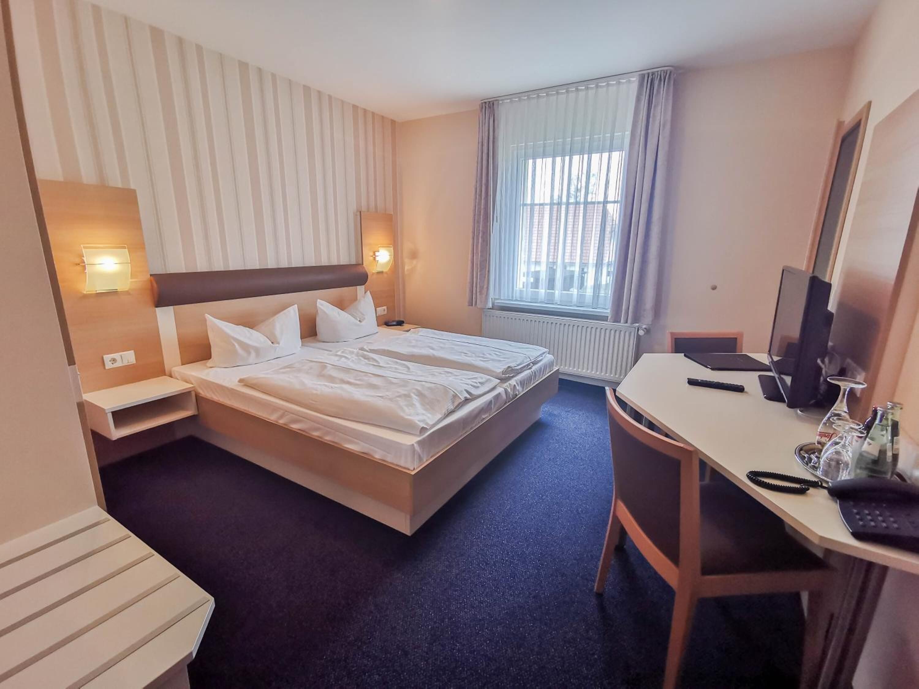 A hotel room with a large bed, two bedside tables, a desk with a chair, a TV, and a window with curtains.
