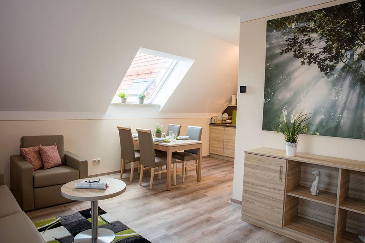 A modern living room with a dining area, a sofa, a round table, and a large window with a view of the sky.