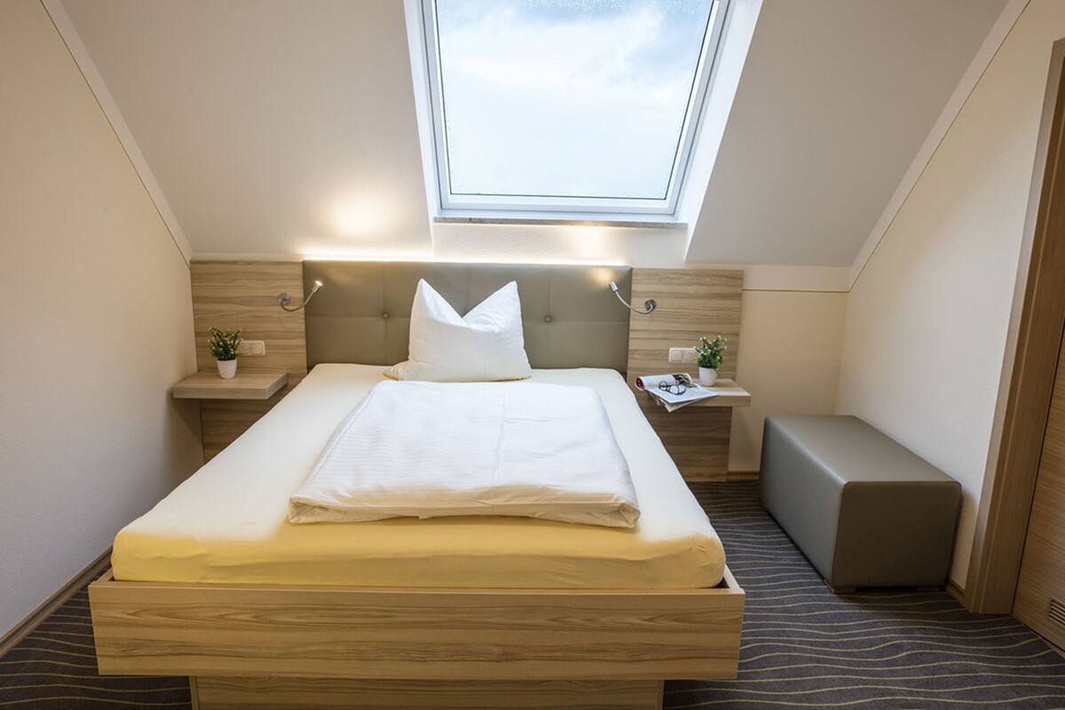 A modern bedroom with a skylight, a bed with white linens, a bedside table with a plant, and a bench.