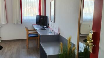 Ein Zimmer mit Schreibtisch, Stuhl und einem großen Spiegel. Ein Fenster mit Vorhängen und ein Heizkörper an der Wand.