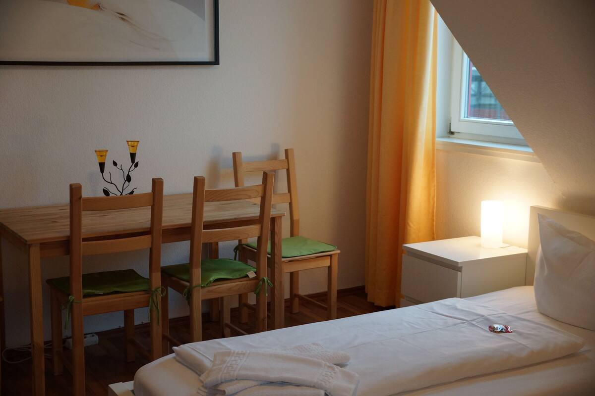 Quarto com cama, escrivaninha, cadeiras, mesa de cabeceira e janela com cortinas.