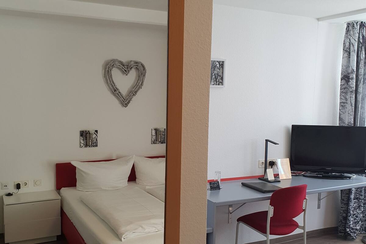 Quarto de hotel com cama, escrivaninha, cadeira, televisão e uma decoração em formato de coração na parede.