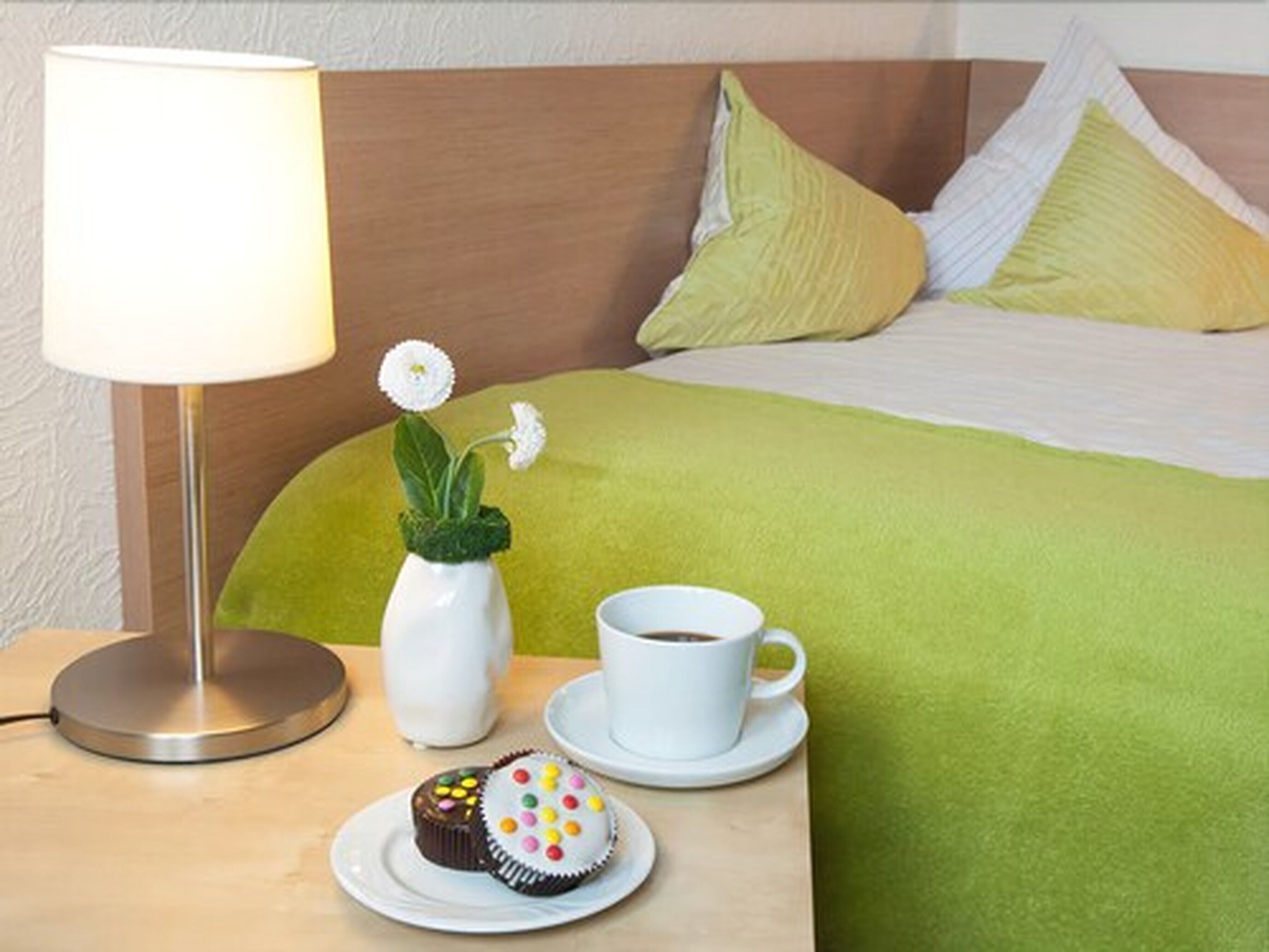 A neatly made bed with a green blanket, a white vase with a white flower, a cup of coffee, and two cupcakes on a small table.
