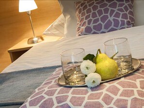 A neatly made bed with a patterned pillow, a tray with two glasses, a pear, and a white flower on a hotel room.