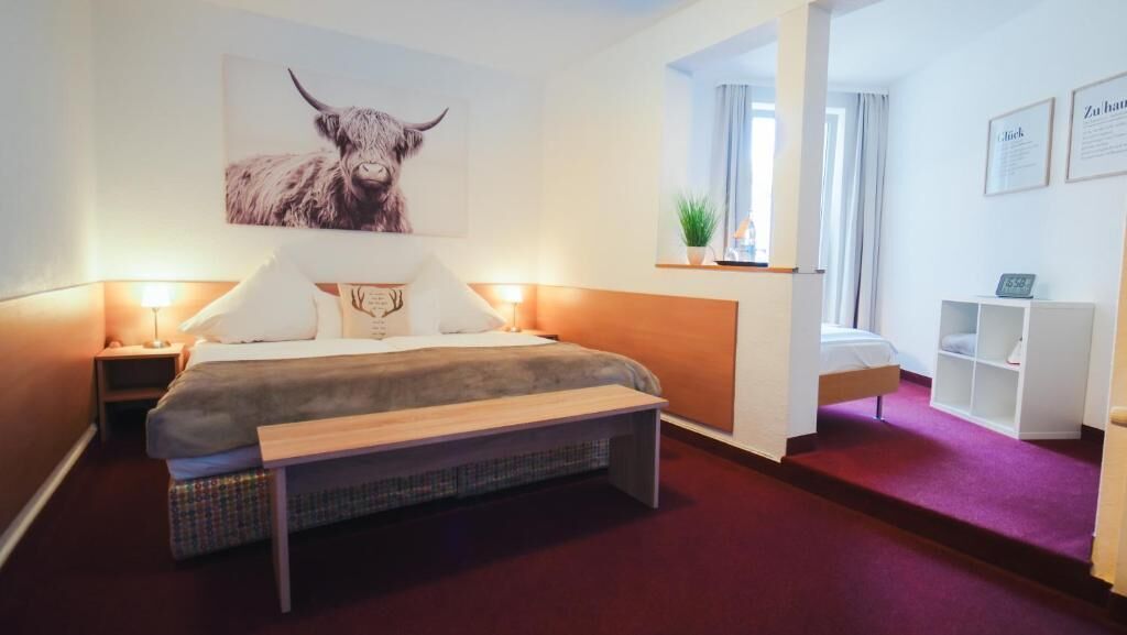 A hotel room with a bed, bedside tables, a bench, a wall-mounted artwork of a bull, a small shelf with a plant, and a window with curtains.