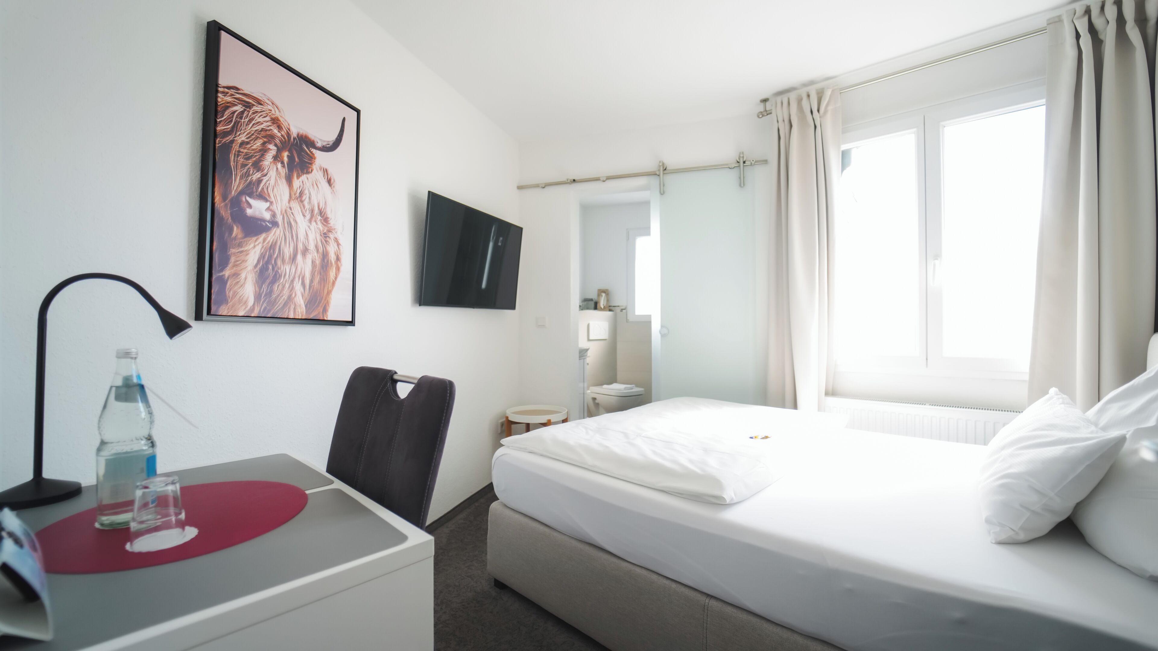 Ein Hotelzimmer mit Bett, Schreibtisch mit Stuhl, Fernseher, Bad und Fenster mit Vorhängen.