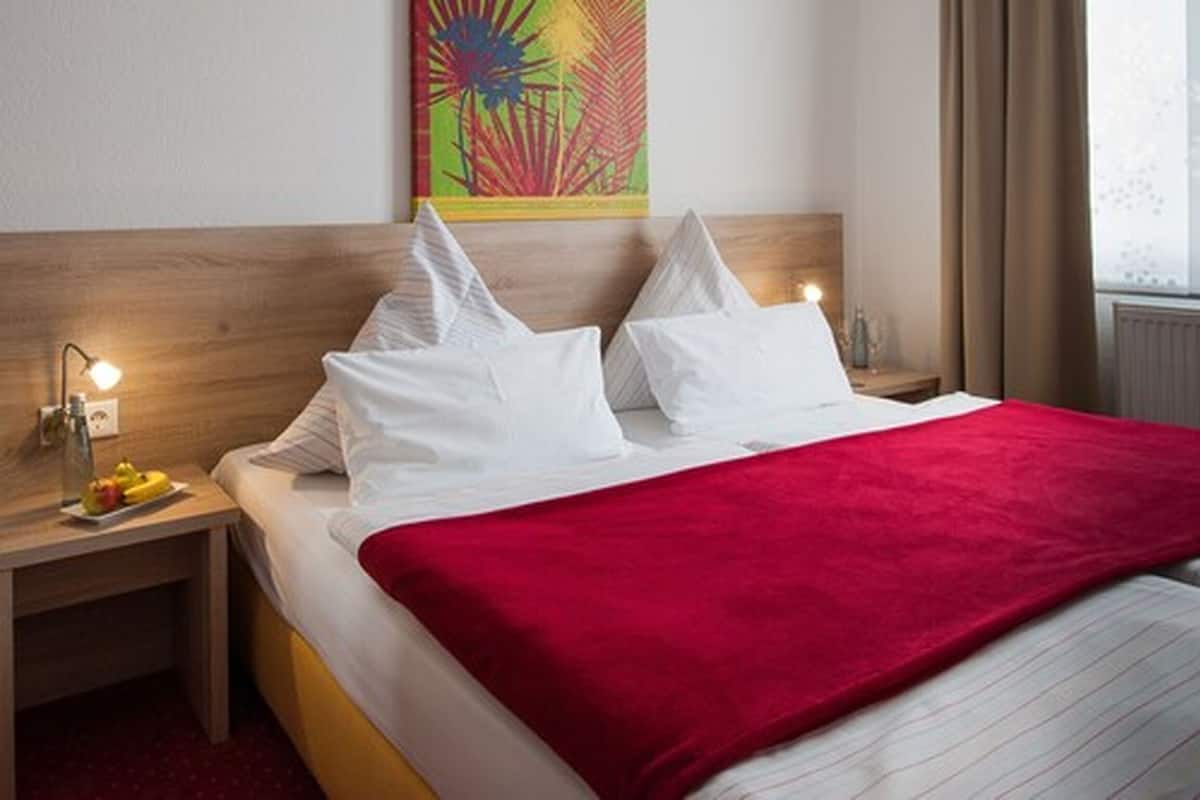 A hotel room with a bed, a red blanket, white pillows, a nightstand with a lamp, and a colorful artwork on the wall.