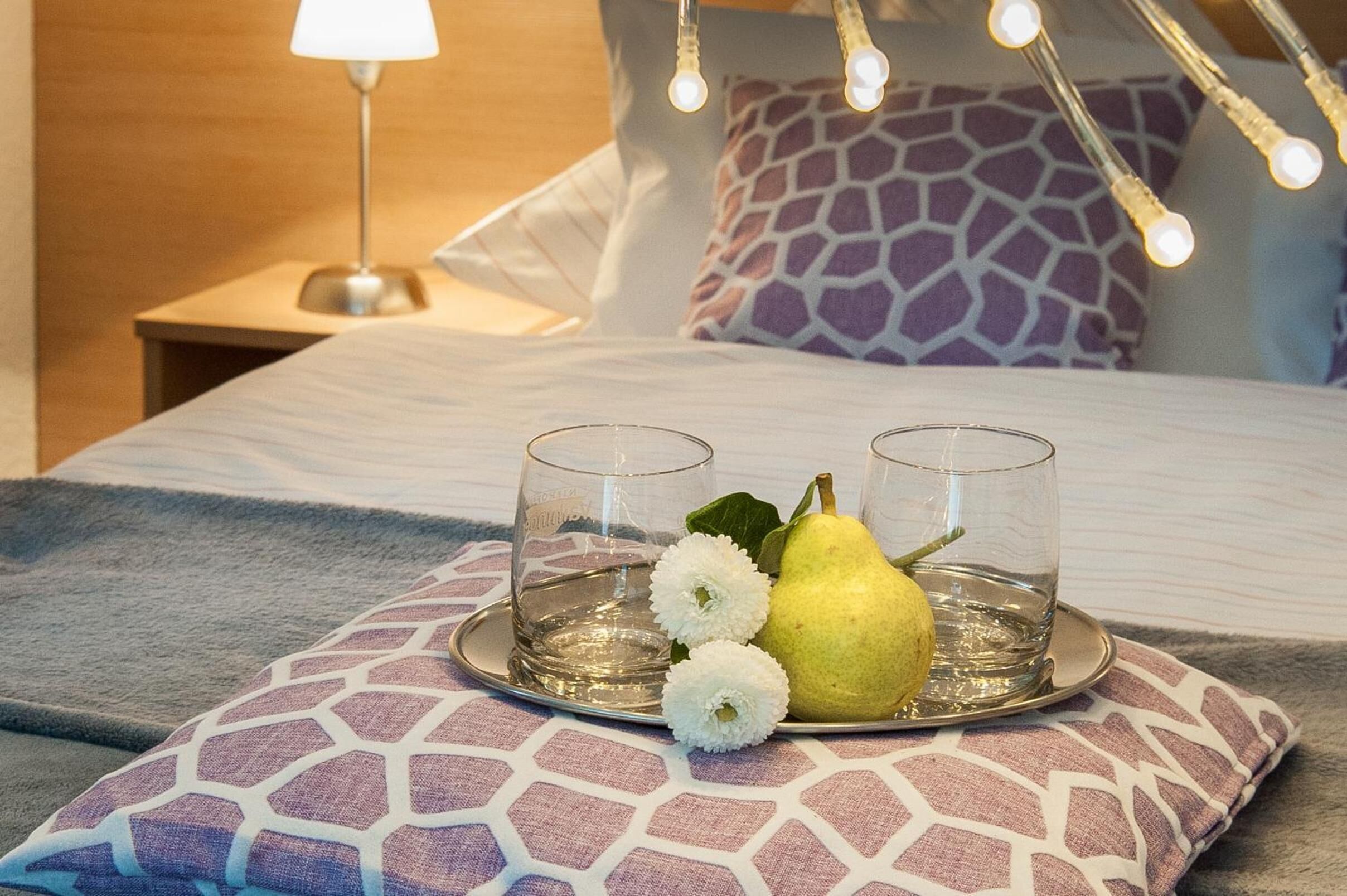 A neatly made bed with a patterned pillow, a tray with two glasses, a pear, and white flowers.