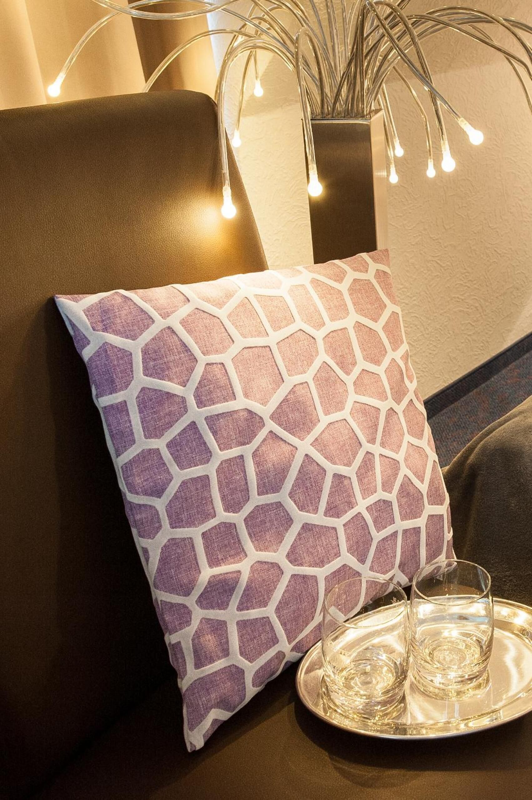 Un sofá con un cojín estampado, una bandeja con vasos y luces de cadena al fondo.