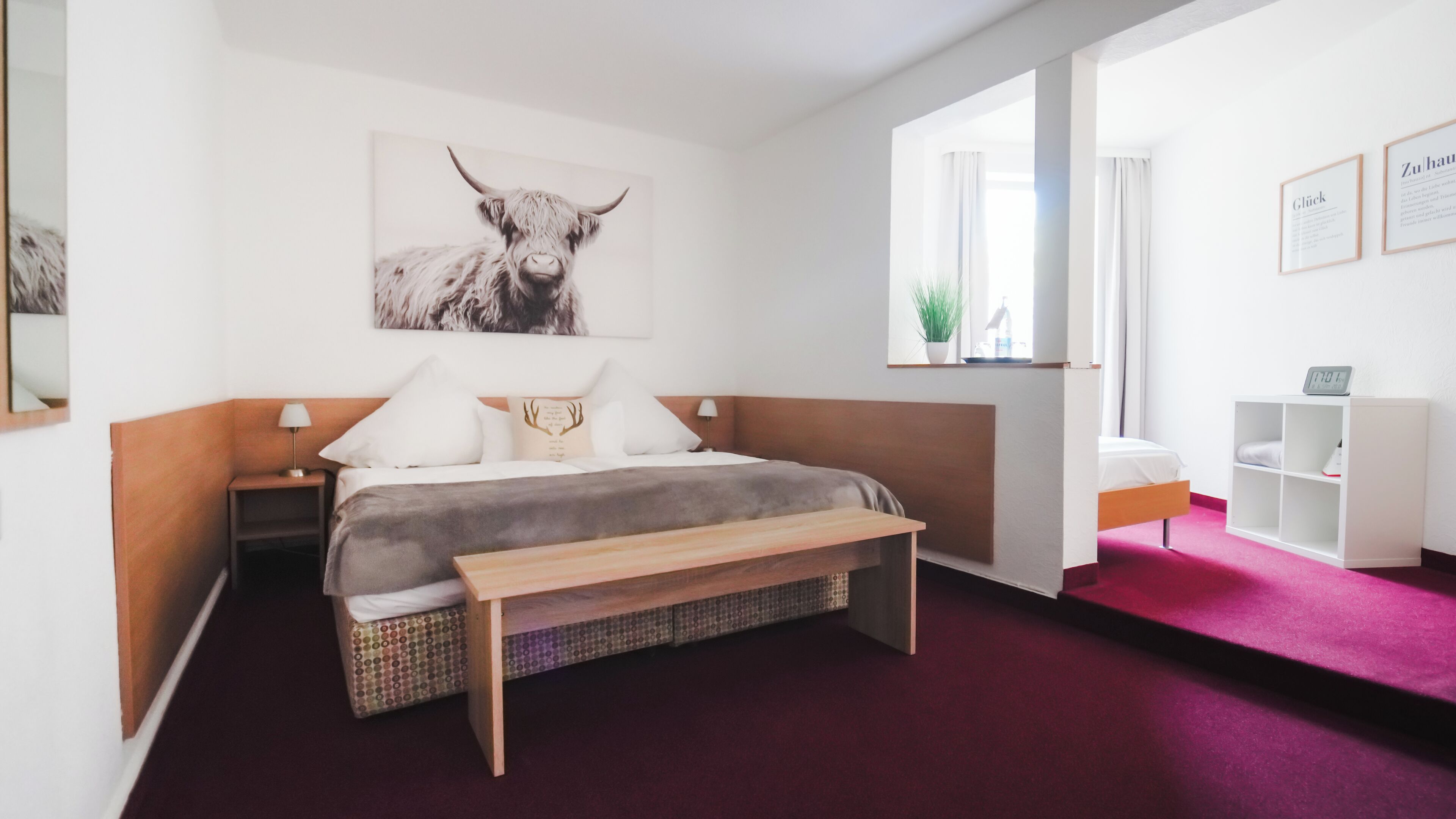 Ein modernes Hotelzimmer mit einem großen Bett, einer hölzernen Bank, einem kleinen Beistelltisch, einer Nachttischlampe, einem gerahmten Kunstwerk mit einem Stier, einer Topfpflanze und einer weißen Regalwand.