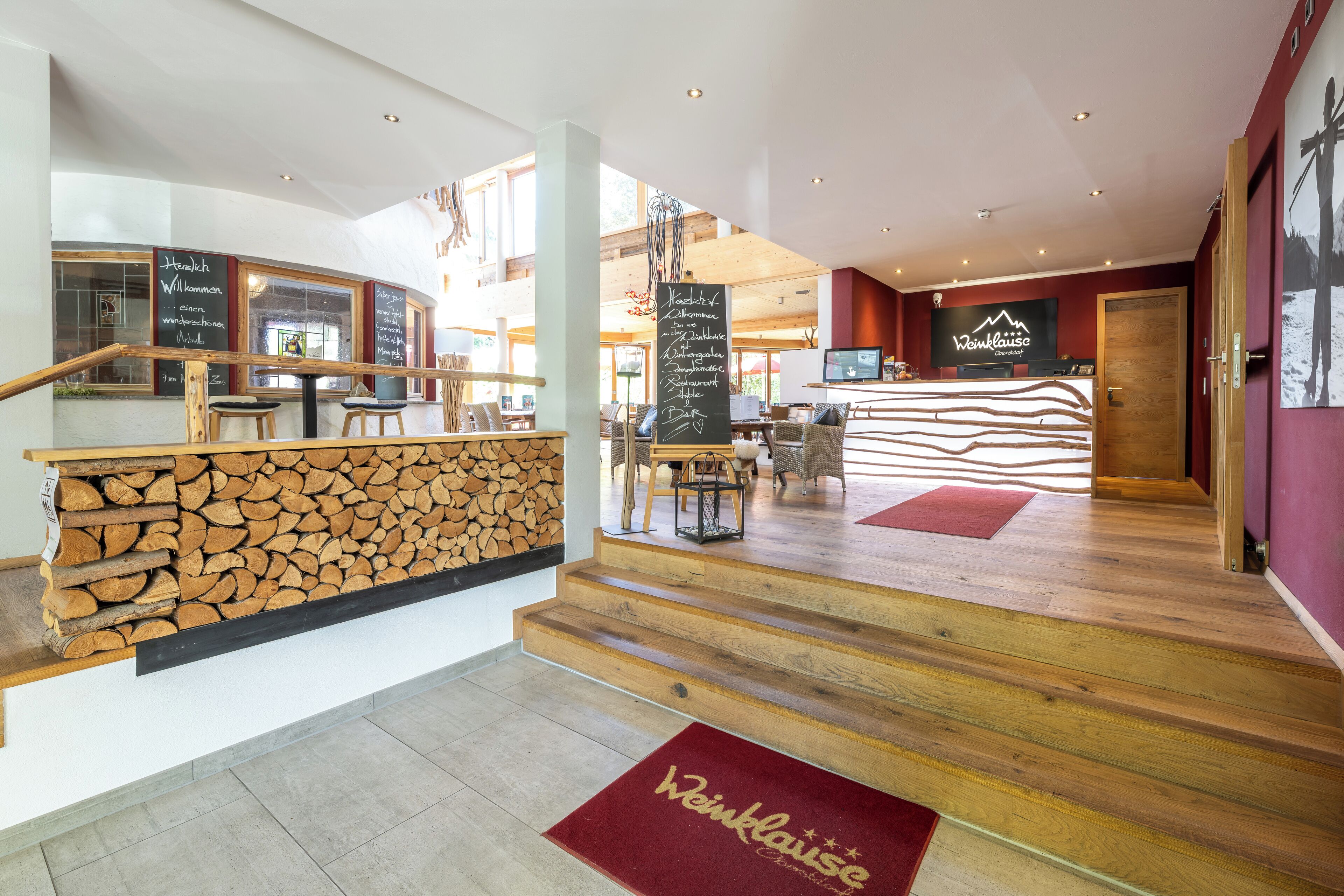 Eine Hotelhalle mit einer Holztreppe, einem Empfangsbereich und einer Sitzecke mit Stühlen und Tischen.