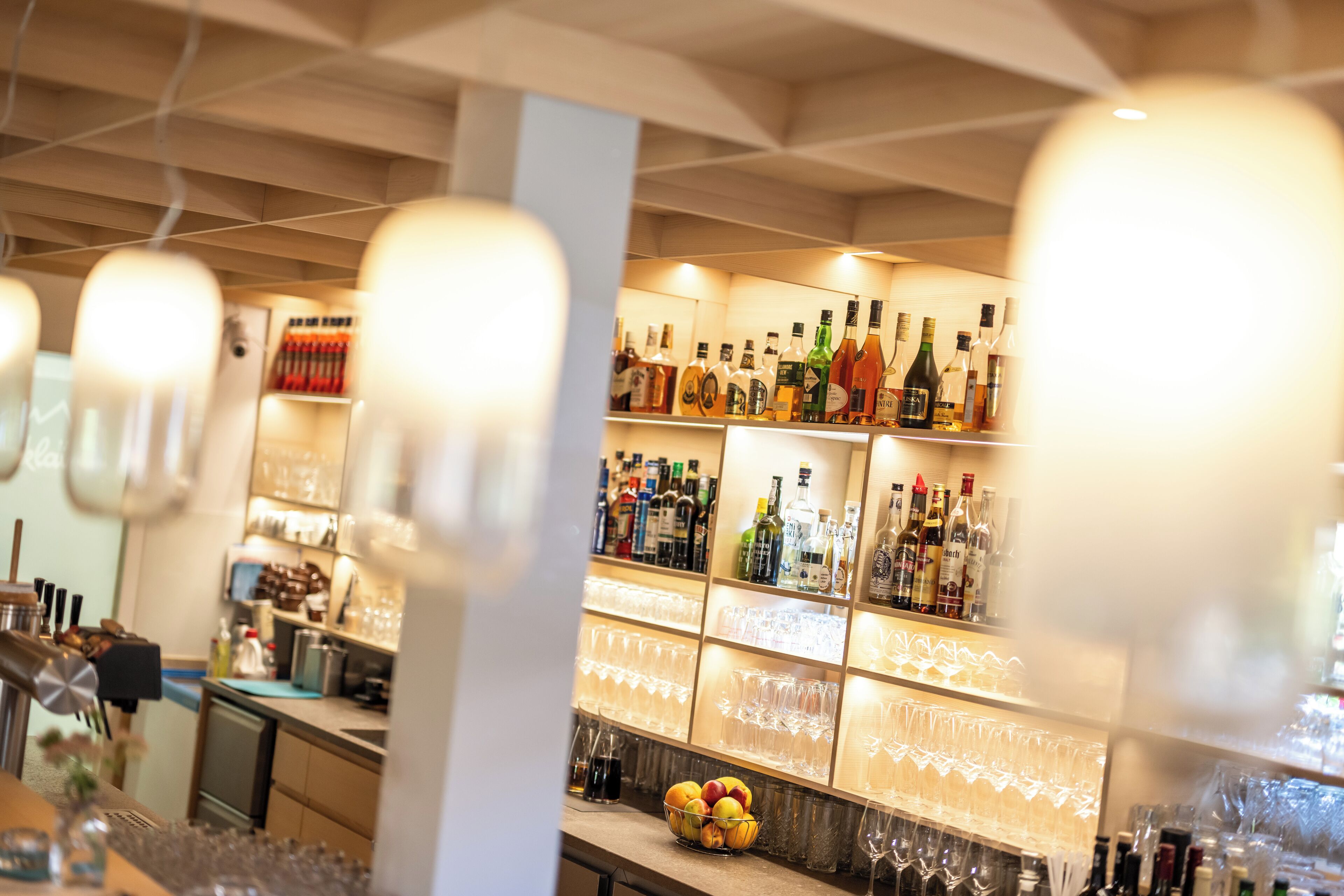 Zona de bar con estantes llenos de botellas de licores y un bol de fruta sobre la barra.