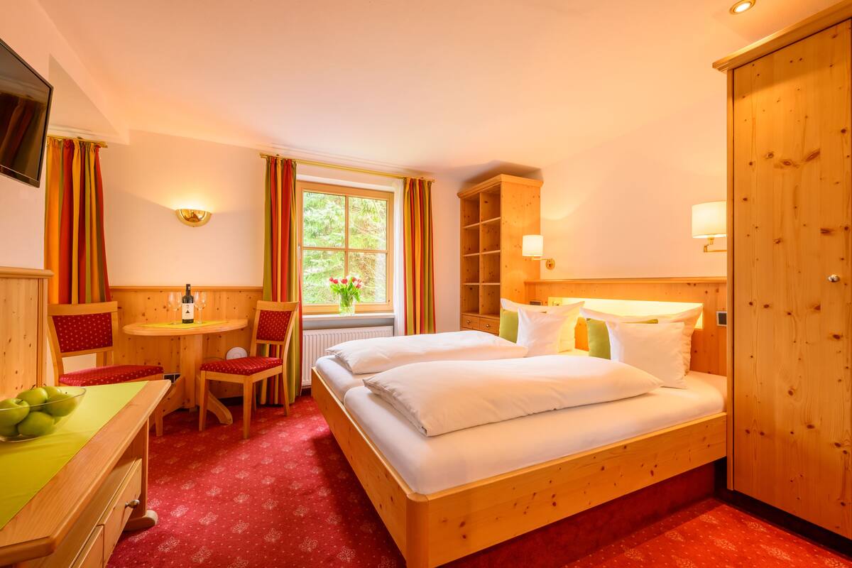 Una camera d'albergo con un letto di legno, una scrivania con una sedia, un tavolino e una finestra con tende.