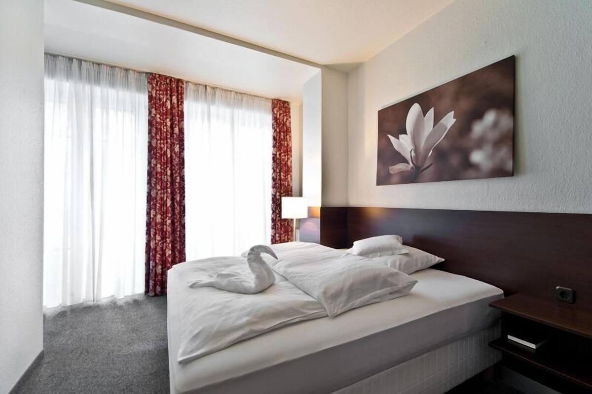Hotellihuone, jossa on suuri sänky, pääty, seinälle ripustettu taulu ja ikkuna, jossa on verhot.