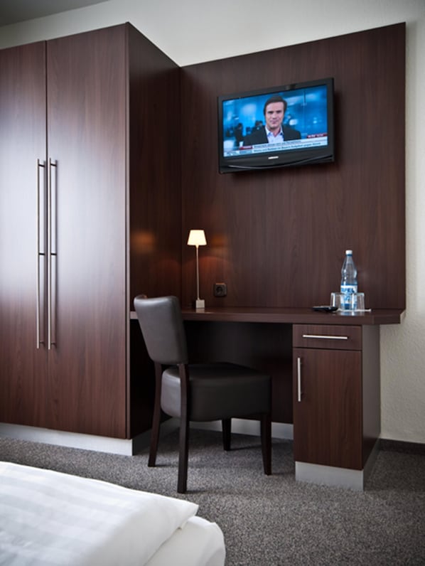 A hotel room with a bed, a desk with a chair, a television mounted on the wall, and a water bottle on the desk.