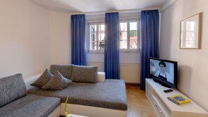 A modern living room with a grey sofa, a television, and a window with blue curtains. - Hotel Schwanen (Mosbach)