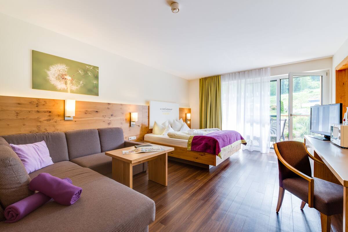 Une chambre d’hôtel moderne avec un canapé, un lit recouvert d’une couverture violette, un bureau en bois et un téléviseur.