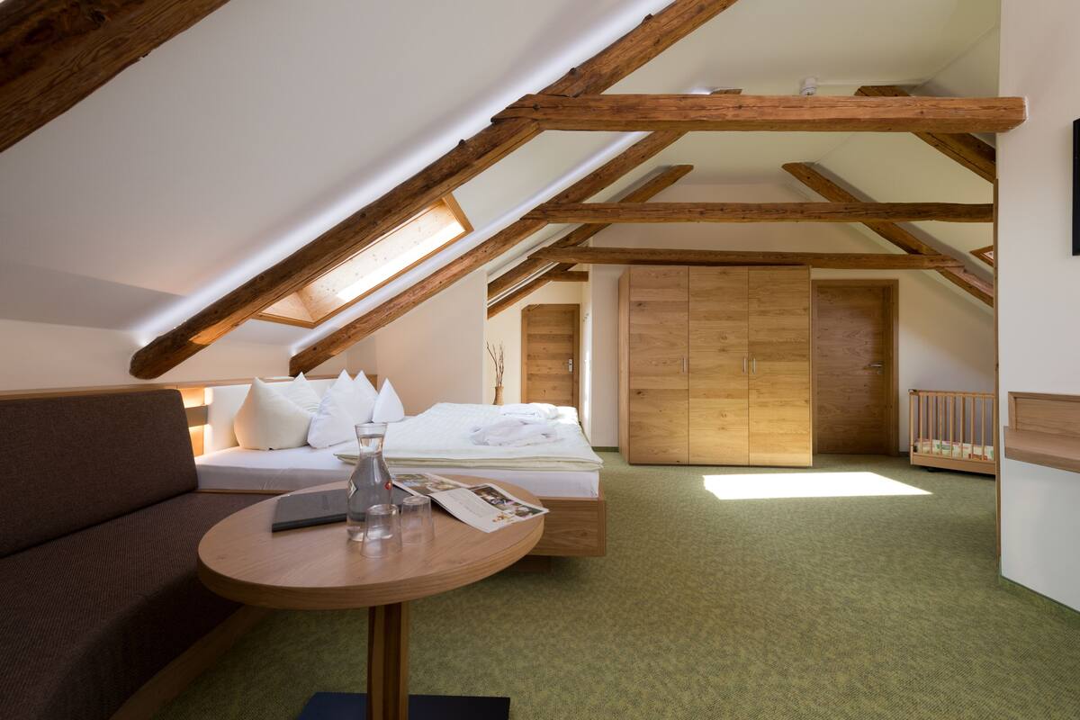 Une pièce avec un plafond en pente, des poutres en bois, un lit avec une literie blanche, un canapé marron, une petite table ronde avec un vase, et une armoire en bois.