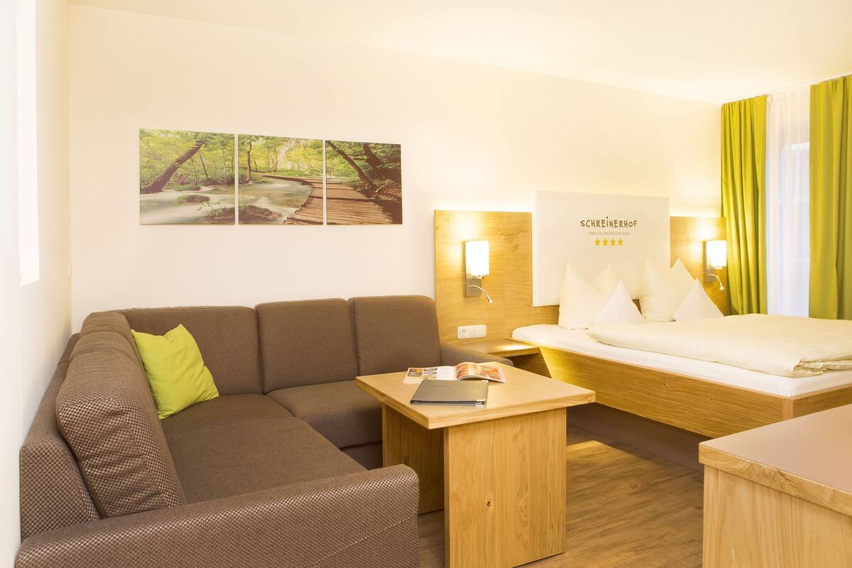 Une chambre d’hôtel avec un canapé, une petite table, un lit et une table de chevet. Il y a une lampe fixée au mur et une affiche accrochée au mur.