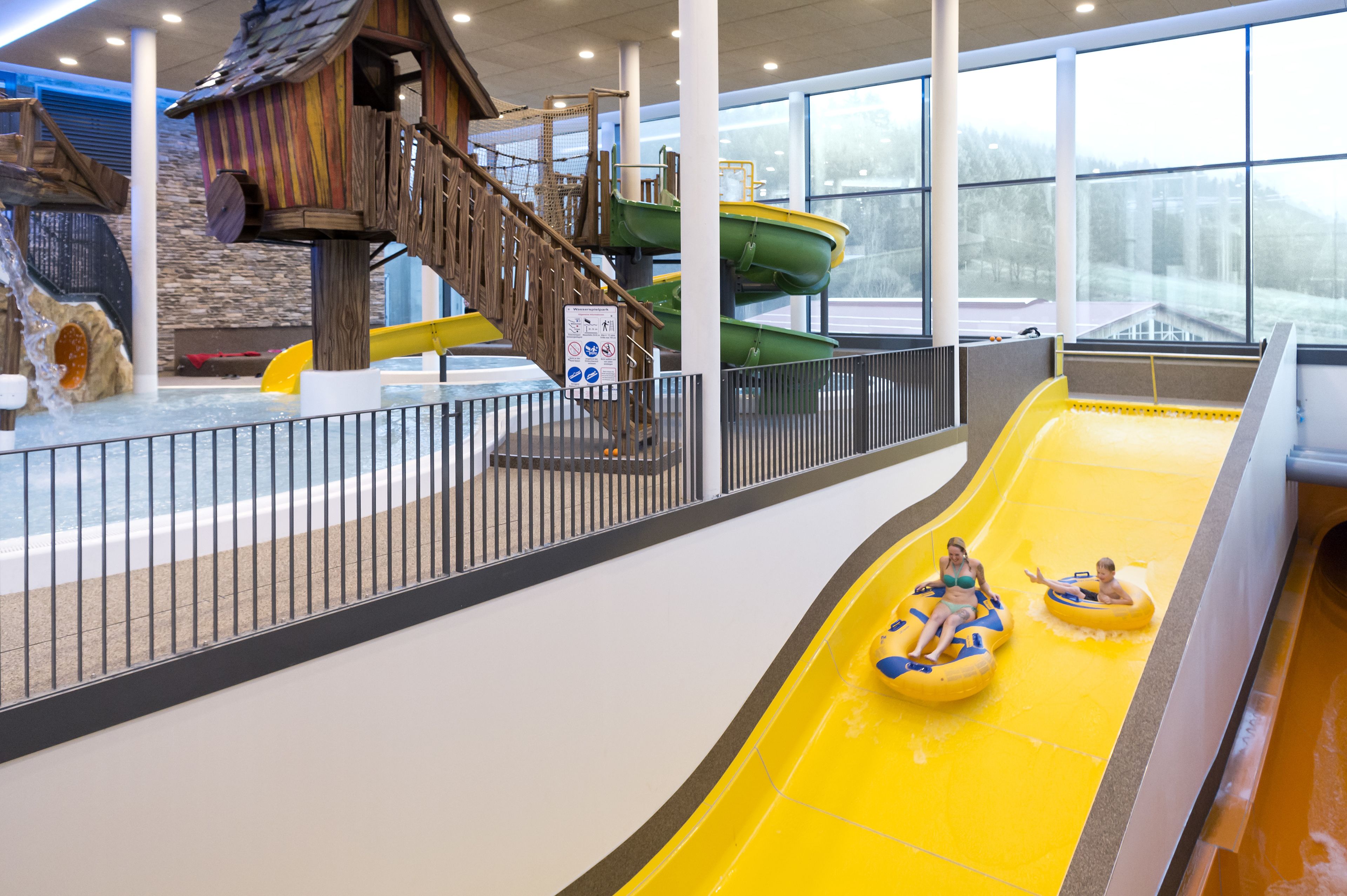 Ein Wasserpark im Innenbereich mit einer gelben Wasserrutsche, einem hölzernen Spielhäuschen und einer grünen Wasserrutsche.