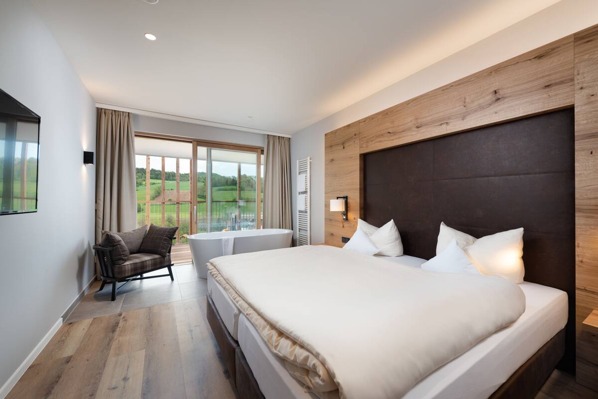 Une chambre d’hôtel moderne dotée d’un grand lit, offrant une vue sur l’extérieur, d’un coin salon avec un fauteuil et d’un mur décoratif en bois.