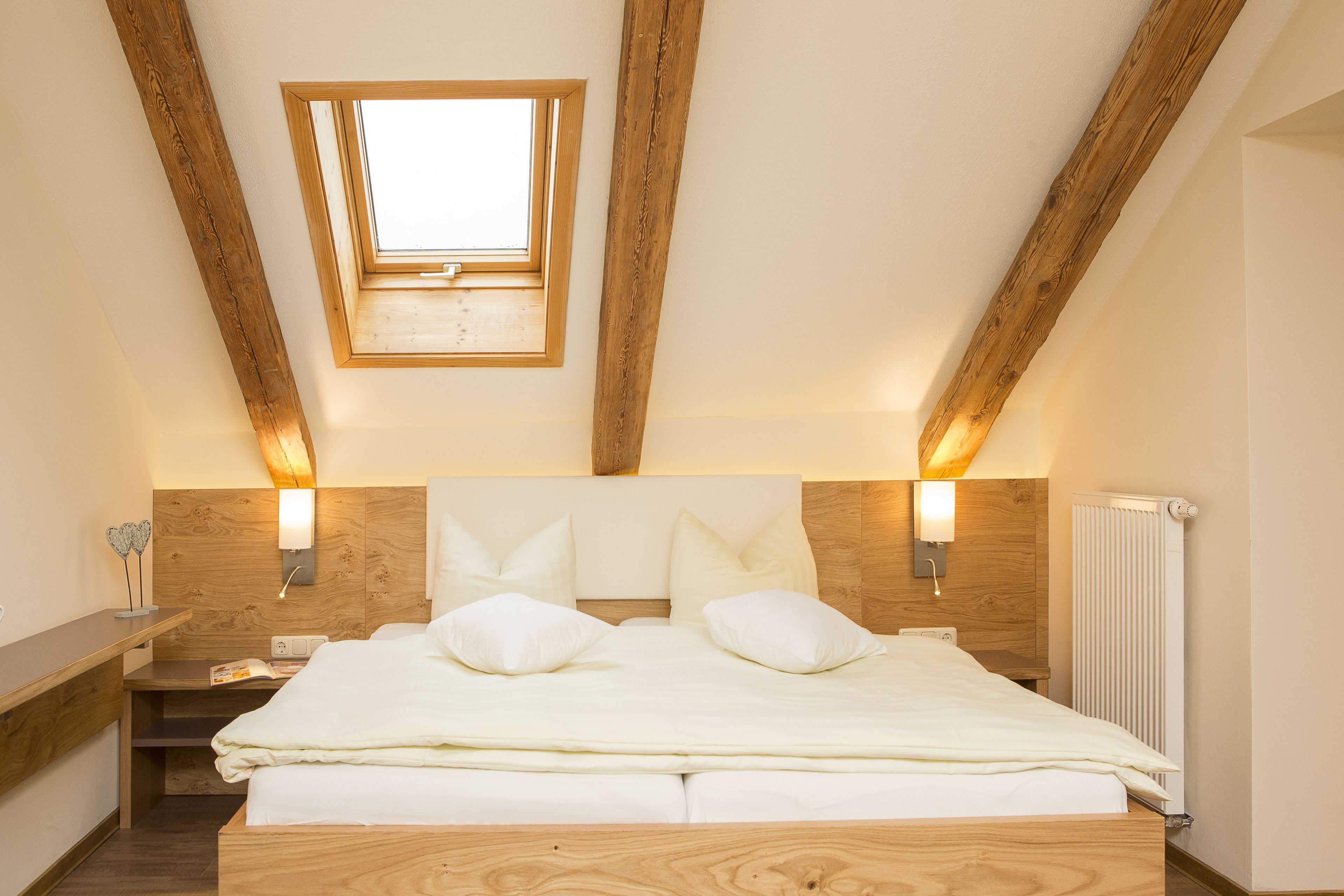 A bedroom with a sloped ceiling, wooden beams, a skylight, a bed with pillows, bedside lamps, and a radiator.