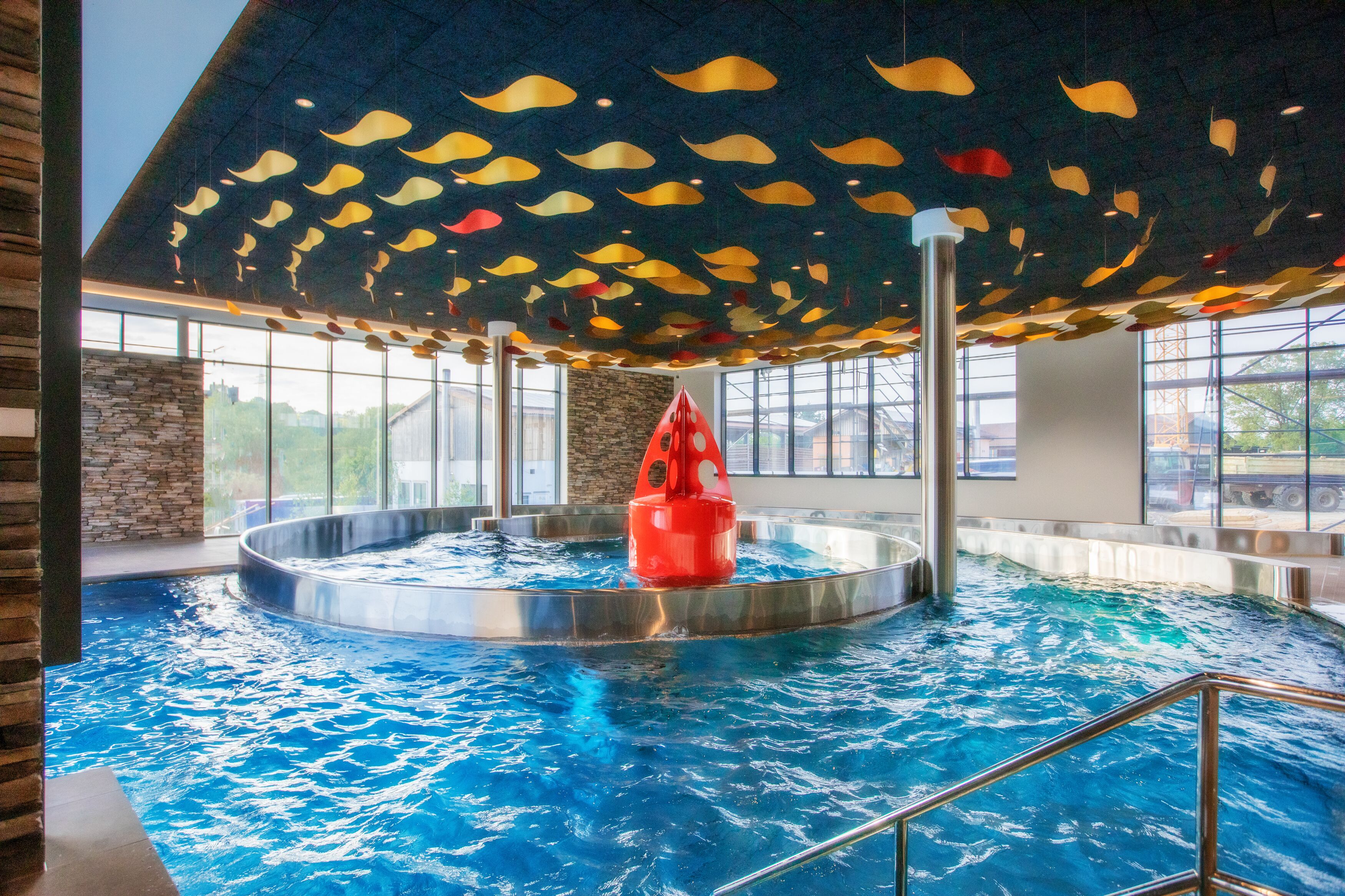 En indendørs swimmingpool med et centralt rødt vandelement, omgivet af store vinduer og et loft med hængende, bladformede dekorationer.