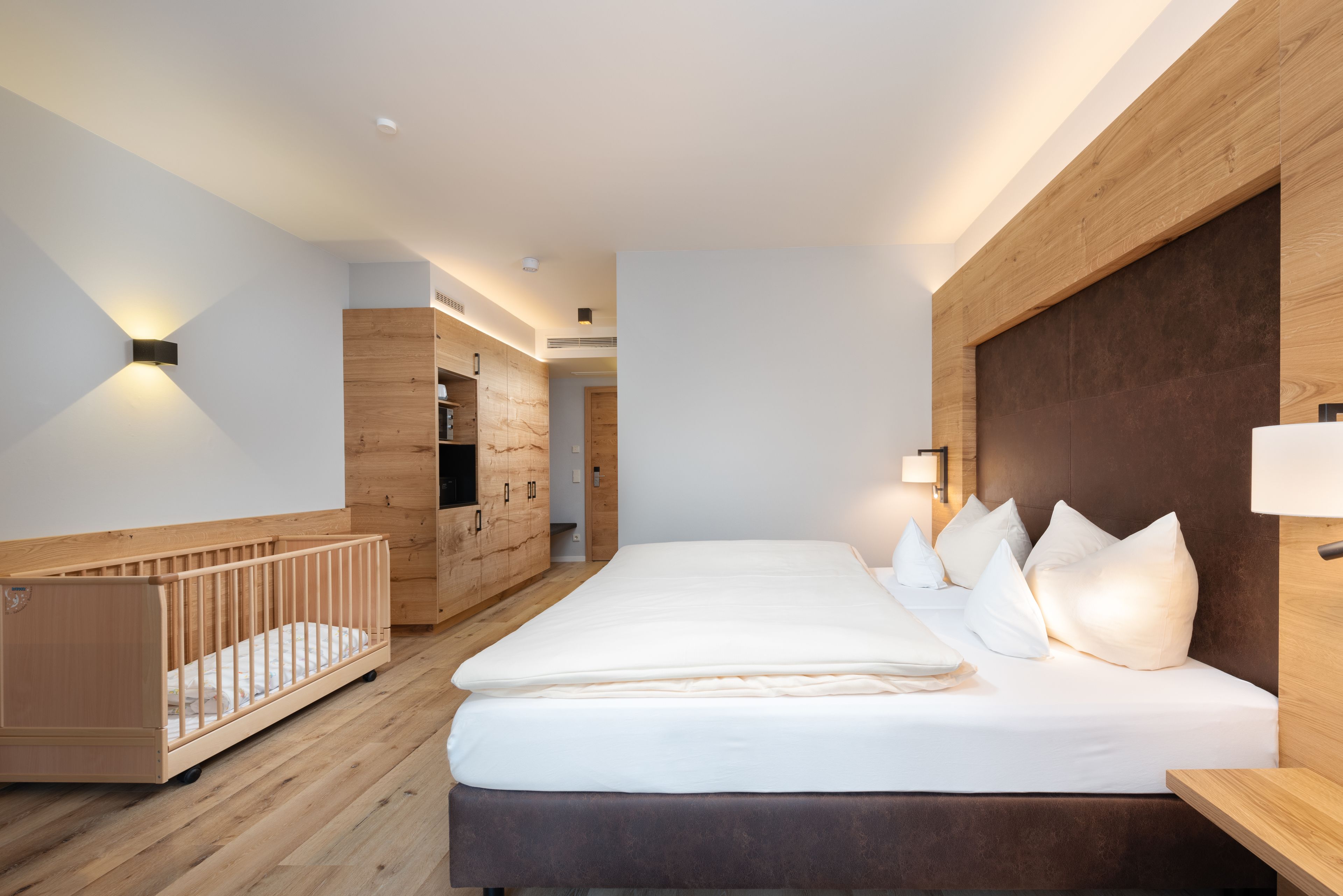 A modern bedroom with a wooden crib, a large bed with white bedding, and a wooden wardrobe.